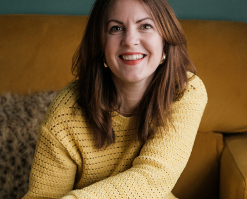Kelly Molson, Agency Advisor smiles at the camera wearing a yellow sweater while sitting on a brown sofa.